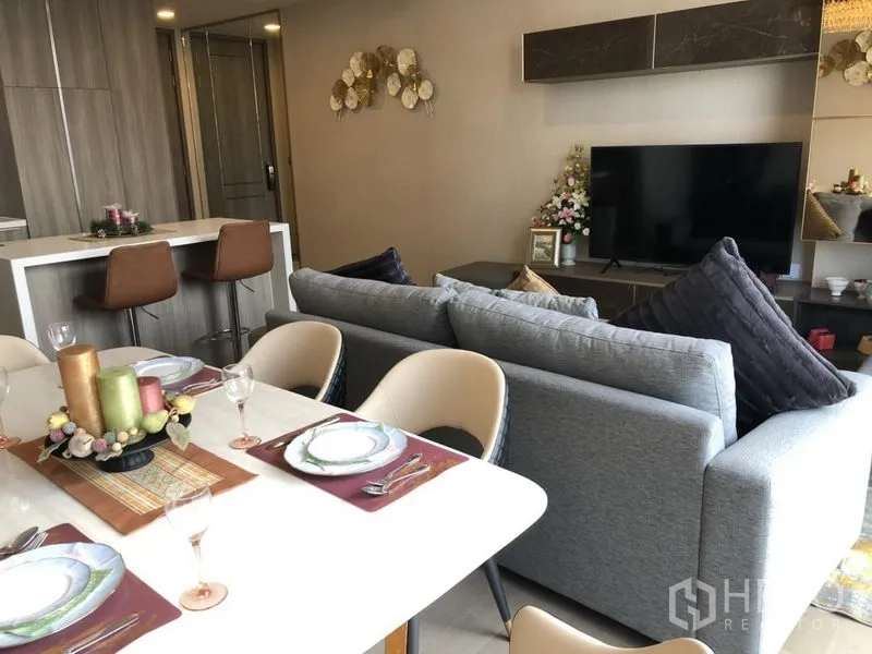 公寓 出租在Watthana, Bangkok - 客餐一体空间，灰色沙发、电视墙与已摆台的餐桌，旁连厨房。