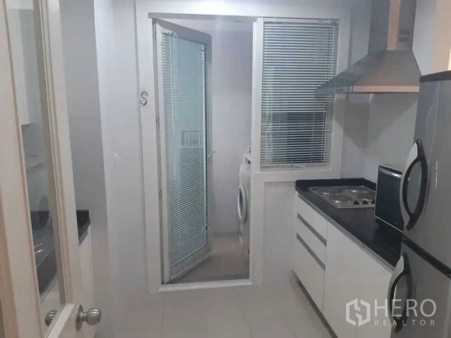 Condominium for rent in Khlong Toei, Bangkok - Bright separate kitchen with black countertops, cooktop, and glass door to a laundry area with washing machine.
