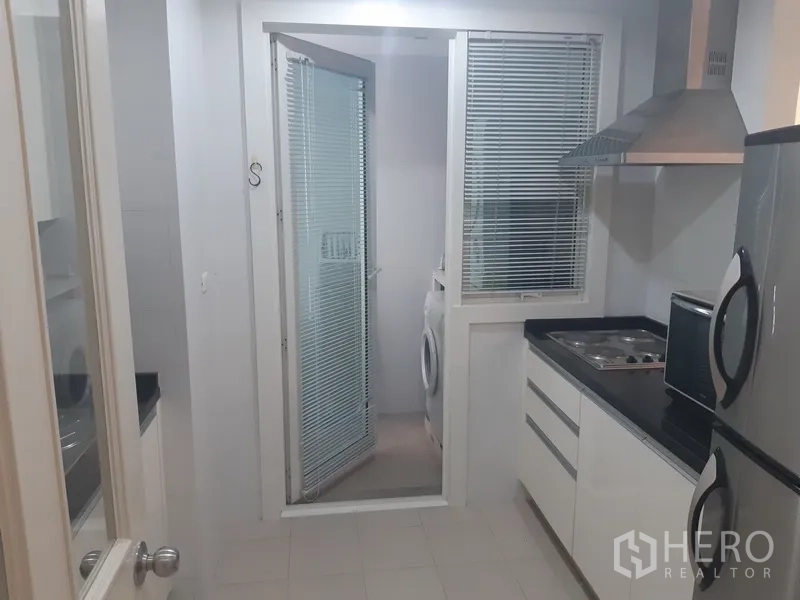 Condominium for rent in Khlong Toei, Bangkok - Bright separate kitchen with black countertops, cooktop, and glass door to a laundry area with washing machine.