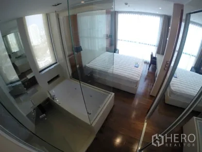 Condominium for rent in Khlong Toei, Bangkok - Master bedroom with floor-to-ceiling windows, hardwood floors, and a glass-walled bathtub suite.