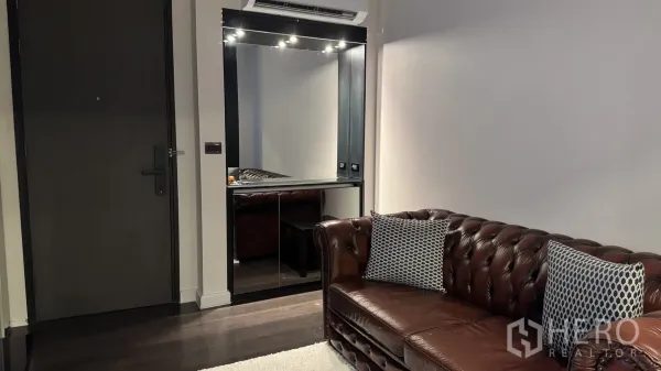 Condominium for rent or sale in Khlong Toei, Bangkok - Entry area with large wall mirror, accent lighting and a brown leather sofa on dark wood flooring.