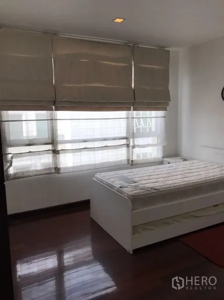 公寓 出租在Watthana, Bangkok - 卧室配深色木地板、罗马帘与白色床架，采光充足的瓦塔那区公寓。