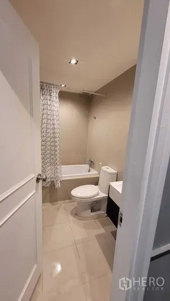 Condominium for rent in Khlong Toei, Bangkok - Modern condo bathroom with bathtub, toilet and compact vanity under warm recessed lighting.