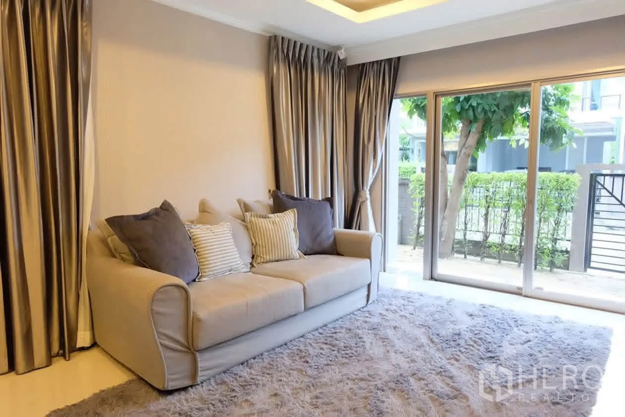 半独立式住宅 for rent or sale in Suan Luang, Bangkok - 客厅配米色沙发与灰色窗帘，大窗临向绿篱与露台景观。
