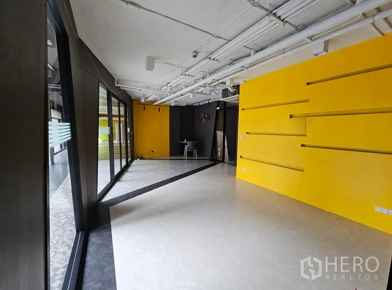 Retail Space for rent - Open-plan retail interior featuring a yellow display wall, shelving, and exposed ceiling pipes.