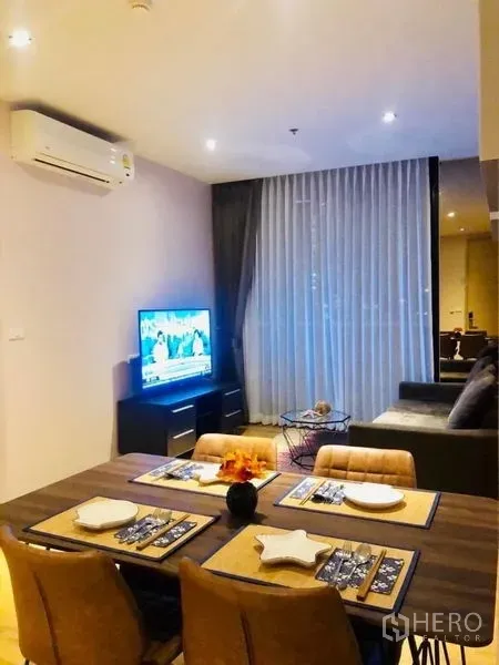 Condominium for rent in Khlong Toei, Bangkok - Dining table for four next to TV console and sofa, with sheer curtains and balcony access.
