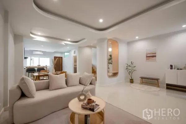 Townhouse for sale in Khlong Toei, Bangkok - Open‑plan living area with sculpted ceilings, soft lighting, beige sofa and dining beyond.