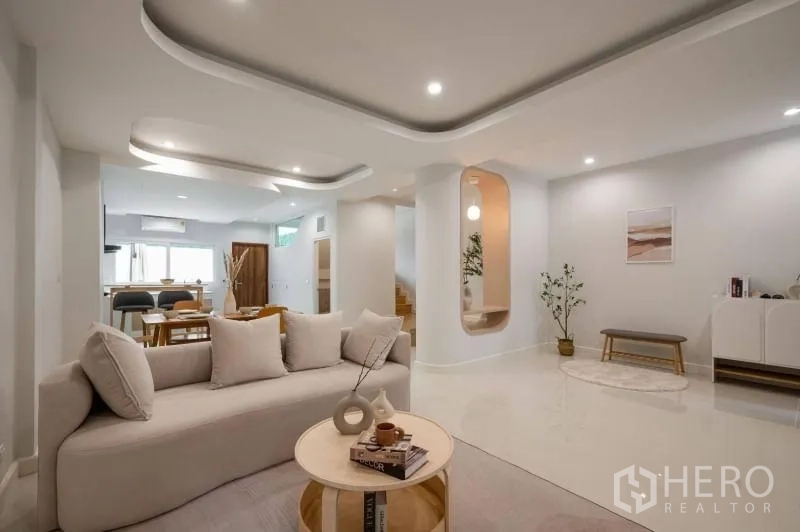 Townhouse for sale in Khlong Toei, Bangkok - Open‑plan living area with sculpted ceilings, soft lighting, beige sofa and dining beyond.