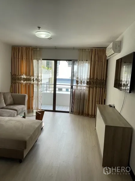 Condominium for rent or sale in Watthana, Bangkok - Living room with wall-mounted TV, air conditioner and sliding doors to the balcony.