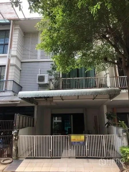Townhouse for sale - Front view of the townhouse showing metal fence, sliding glass doors, balcony and awning shaded by a mature tree.