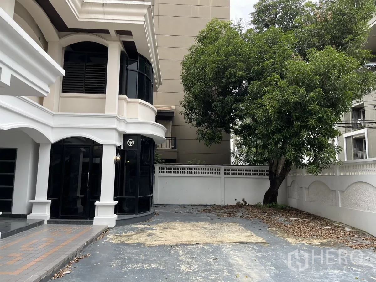 Detached House for rent in Watthana, Bangkok - Side exterior showing corner glass windows, a wide driveway and a large shade tree.