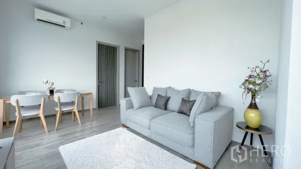 公寓 出租在Watthana, Bangkok - 极简客厅，灰色沙发、空调、用餐角以及通往两间卧室的门。