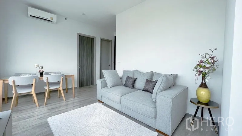 公寓 出租在Watthana, Bangkok - 极简客厅，灰色沙发、空调、用餐角以及通往两间卧室的门。