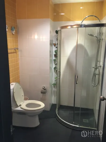 Condominium for rent in Khlong Toei, Bangkok - Bathroom with yellow-and-white tiles, toilet, and curved glass shower enclosure.