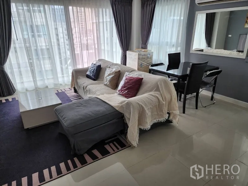 Condominium for rent in Watthana, Bangkok - Sunlit living and dining area with large corner windows, sofa with ottoman, and glass table.