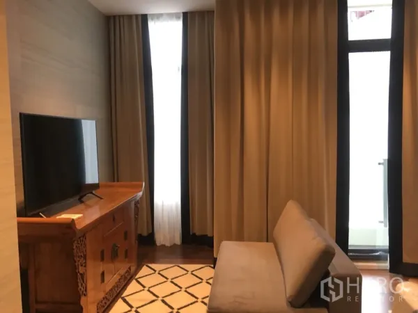 公寓 出租在Watthana, Bangkok - 客厅配木质电视柜、沙发与几何地毯，落地窗帘通向阳台，氛围温馨。
