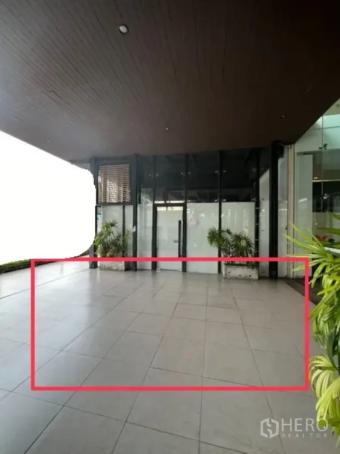 零售空间 出租 - 带大面积玻璃幕墙与瓷砖地面的半室外零售前场，入口处摆放绿植，位于Ekkamai大楼。