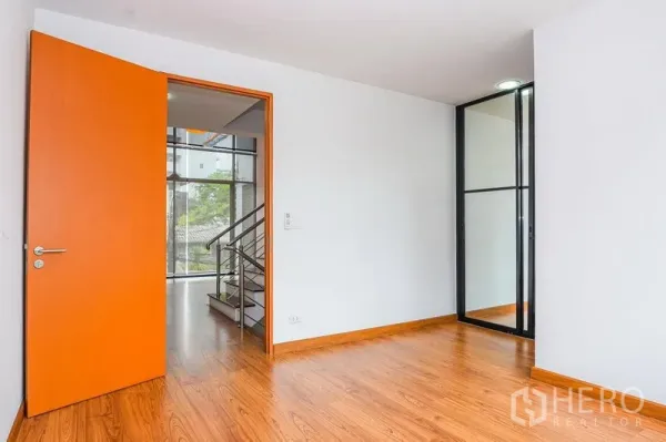 Townhouse for rent in Watthana, Bangkok - Bedroom with wood flooring, orange door and a glass partition for storage or study nook.