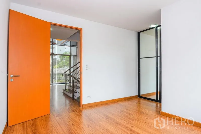 Townhouse for rent in Watthana, Bangkok - Bedroom with wood flooring, orange door and a glass partition for storage or study nook.
