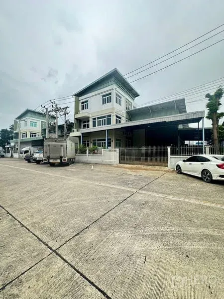 Warehouse for rent or sale in Lam Luk Ka, Pathum Thani - Wide street perspective of the gated warehouse compound and neighboring buildings.