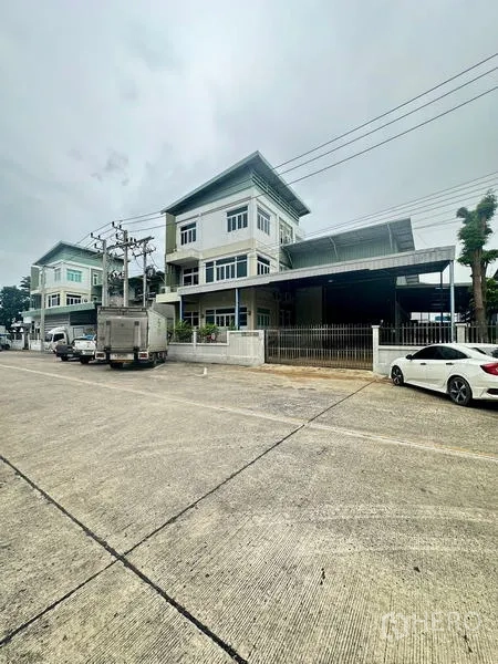 Warehouse for rent or sale in Lam Luk Ka, Pathum Thani - Wide street perspective of the gated warehouse compound and neighboring buildings.