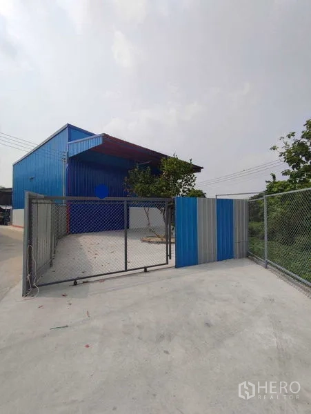 货栈 出租在Pak Kret, Nonthaburi - 带链网围栏的滑动大门，通往仓库庭院与停车区。