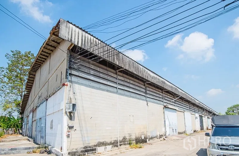 Warehouse for rent in Bang Phli, Samut Prakan - Long side view of a concrete warehouse block with metal roof panels and overhead utility lines under a blue sky.