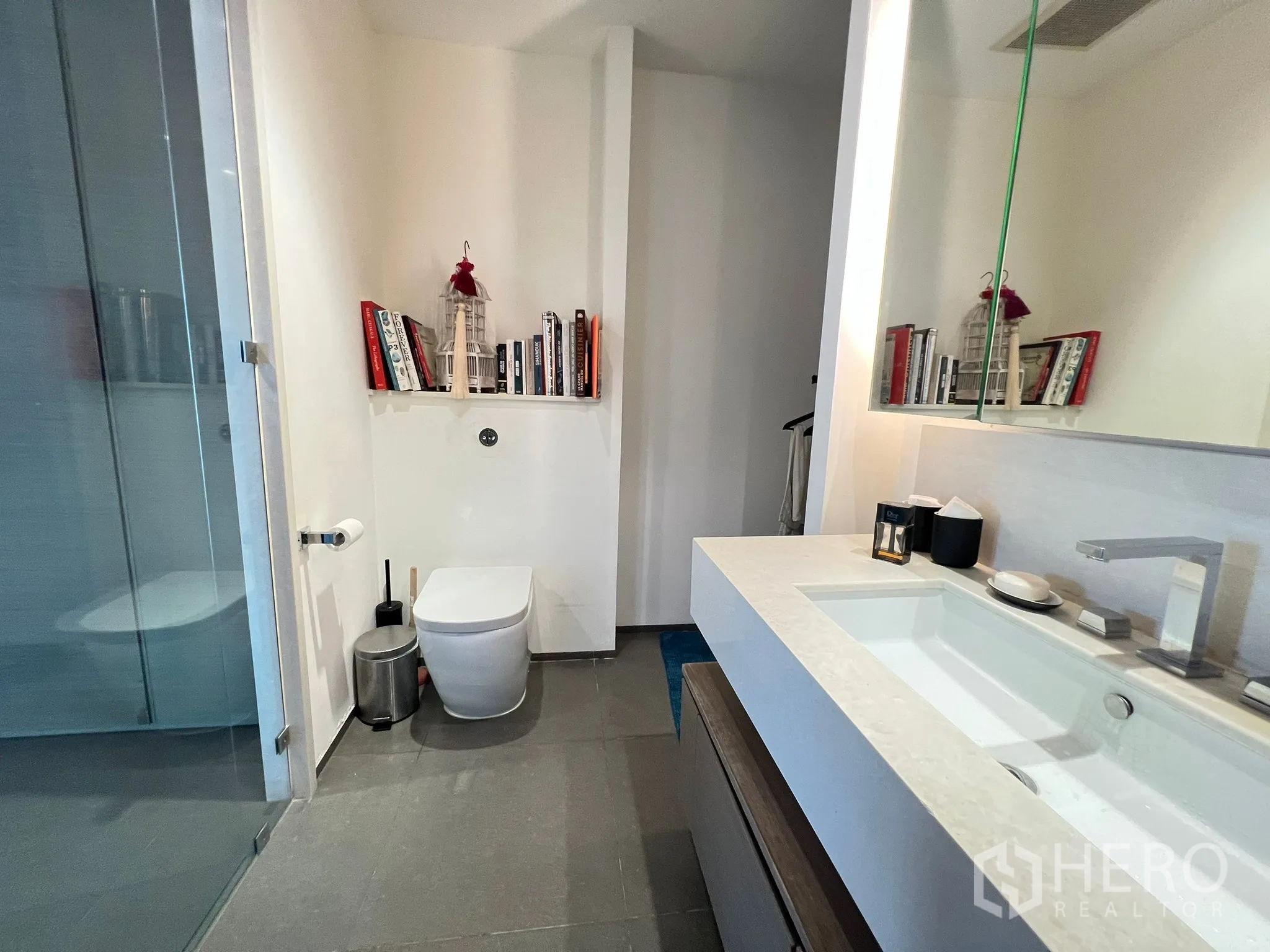 Condominium for sale in Khlong San, Bangkok - Bathroom with large counter-top sink, glass shower, and shelving niche of books.