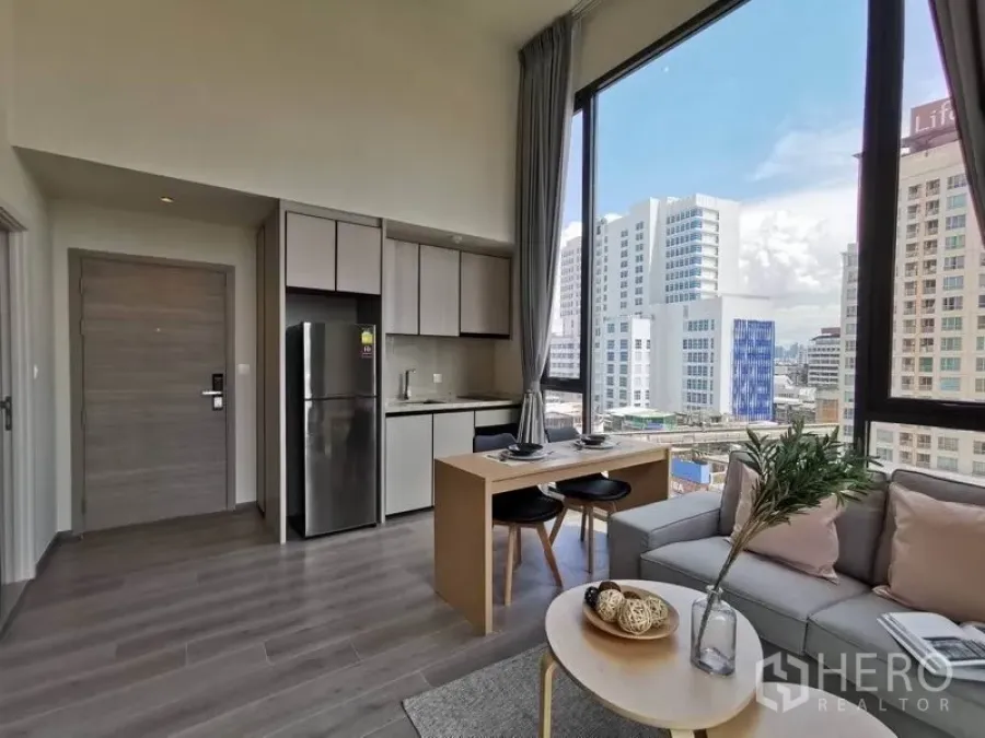 公寓 出租在Phaya Thai, Bangkok - 明亮客厅，带开放式厨房、双人餐桌、沙发与落地窗城市景观。