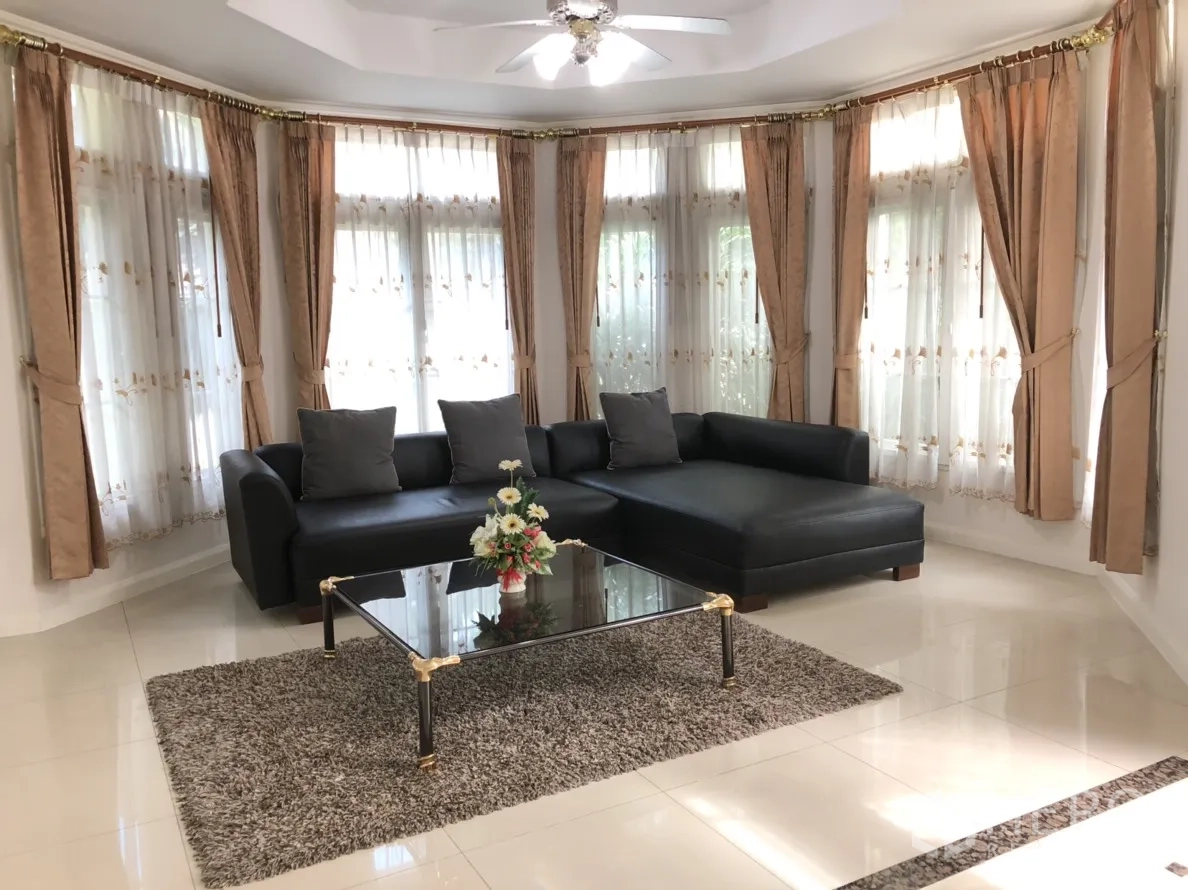 Detached House for rent or sale in Lat Phrao, Bangkok - Bright living room with black L-shaped sofa, glass coffee table, bay windows and ceiling fan.