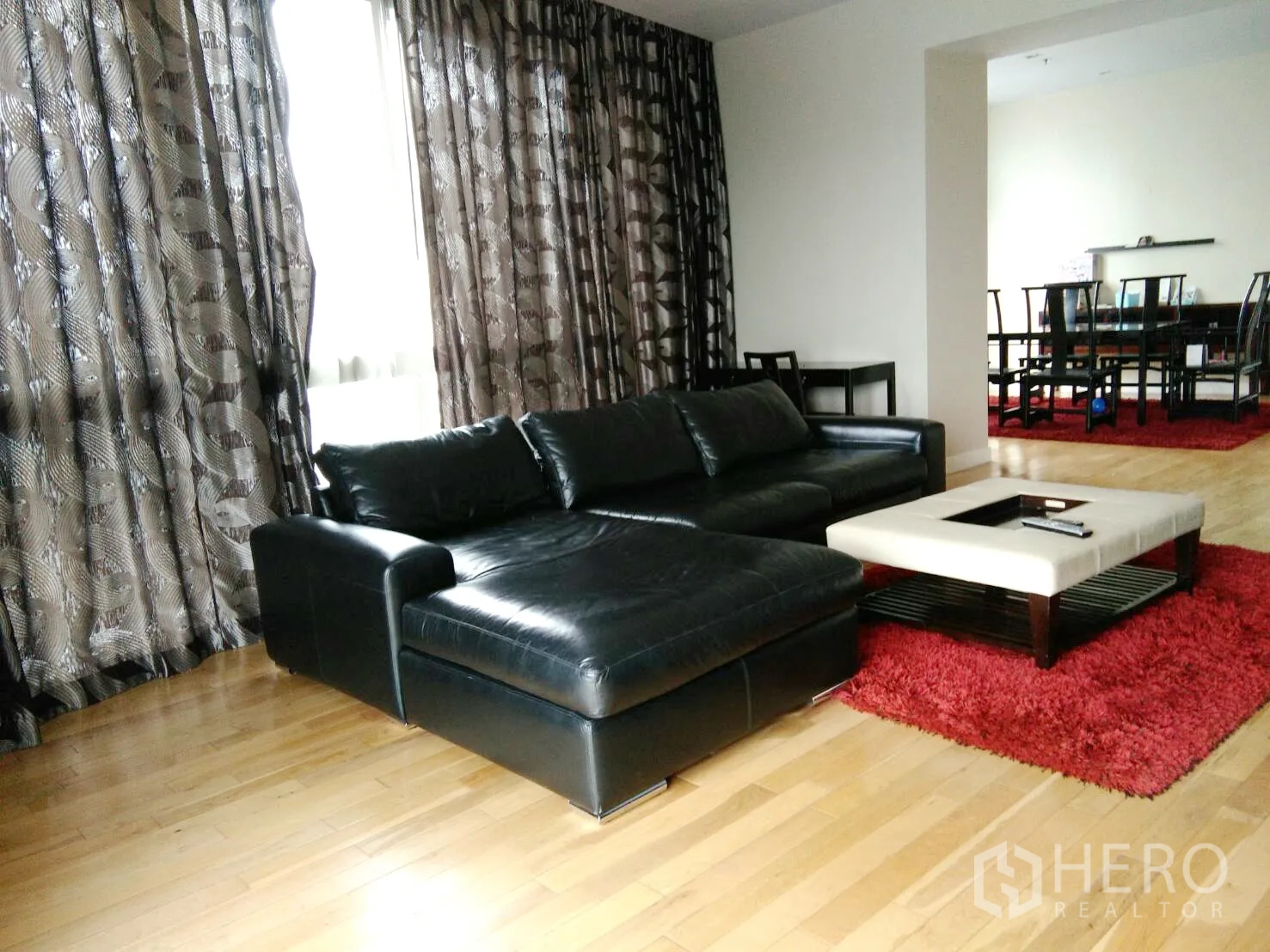 Condominium for rent in Khlong Toei, Bangkok - Spacious living room with black leather sectional sofa and large patterned drapes.