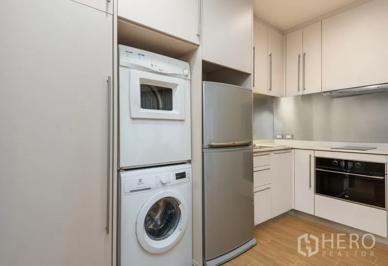 Condominium for rent in Bang Kho Laem, Bangkok - Compact kitchen with white cabinetry, stacked washer and dryer, stainless refrigerator, built-in oven, and electric cooktop.