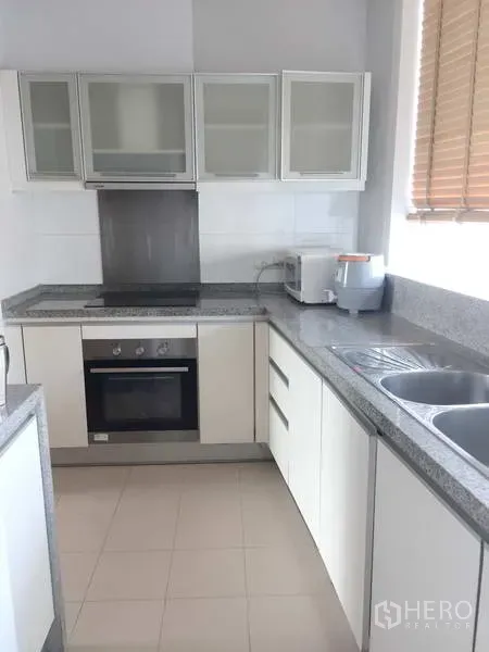 Condominium for sale in Khlong Toei, Bangkok - Modern L-shaped kitchen with granite countertops, electric stove, oven and double sink by a window.