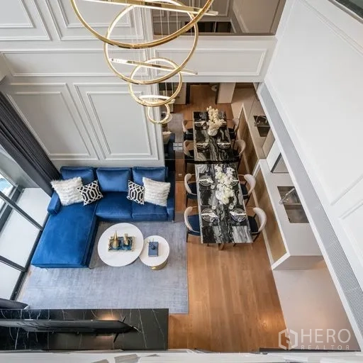 Condominium for rent in Khlong Toei, Bangkok - Overhead view of a double-height living and dining area with a blue sofa, marble dining table, wood floors and a modern gold chandelier.