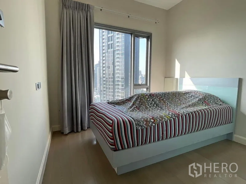 Condominium for sale in Khlong Toei, Bangkok - Cozy bedroom with city-view window, striped bed and gray curtains.