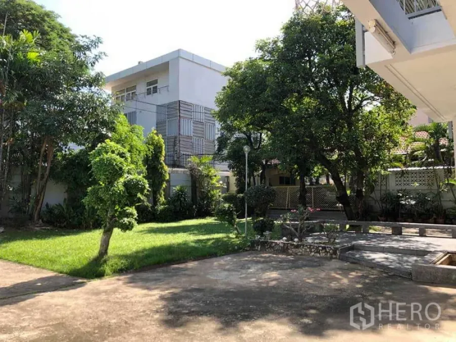 Detached House for rent in Phra Khanong, Bangkok - Shaded front garden with mature trees, lawn and a paved patio of a two‑storey detached house.
