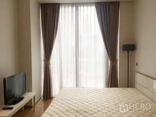 Condominium for rent in Bang Rak, Bangkok - Sunlit bedroom with large sheer-draped windows, a queen bed, TV stand, and floor lamp.
