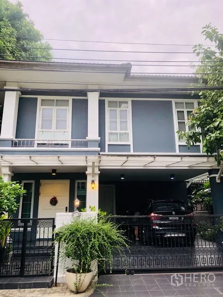 独立式住宅 出租在Bang Kapi, Bangkok - 两层独栋住宅，蓝灰色外立面配白色线条，带围栏门庭与有顶车位，绿意环绕。