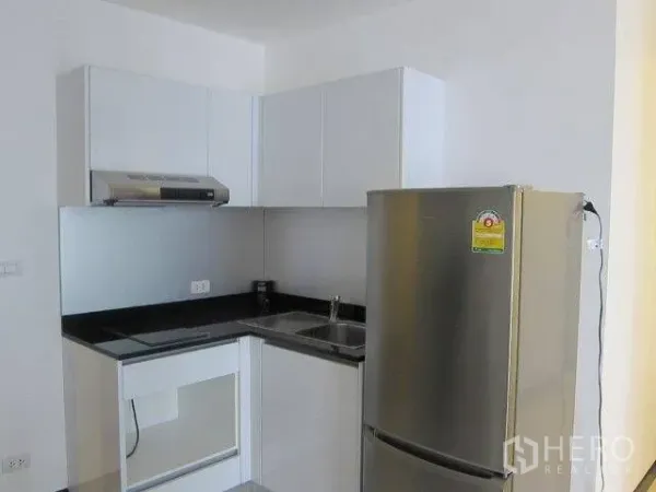 Condominium for rent in Khlong Toei, Bangkok - L-shaped kitchen with white cabinetry, black countertop, sink, range hood, and stainless refrigerator.
