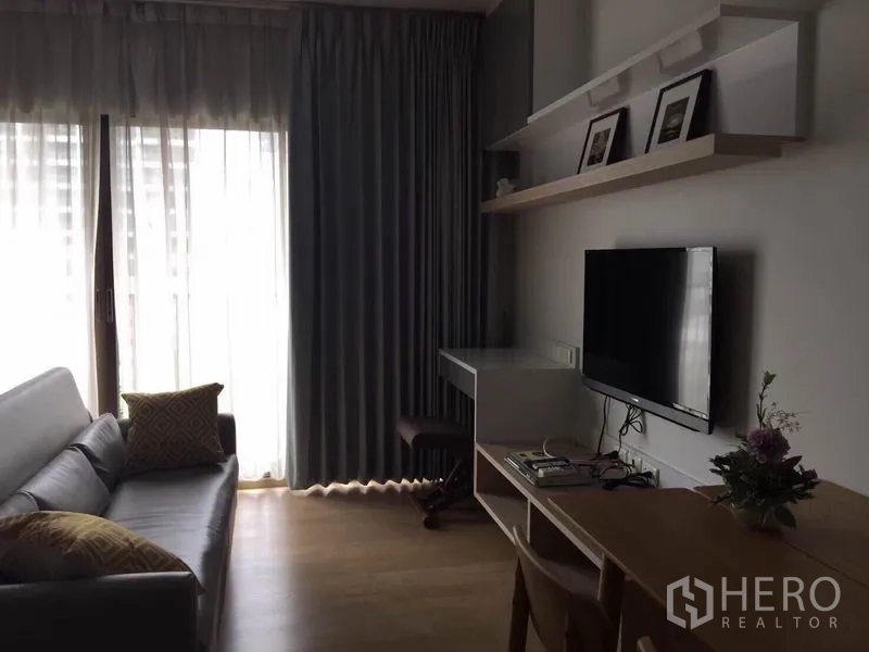 Condominium for rent or sale in Bangkok - Living room with sofa, wall-mounted TV, compact work desk and floor-to-ceiling curtains filtering natural light.