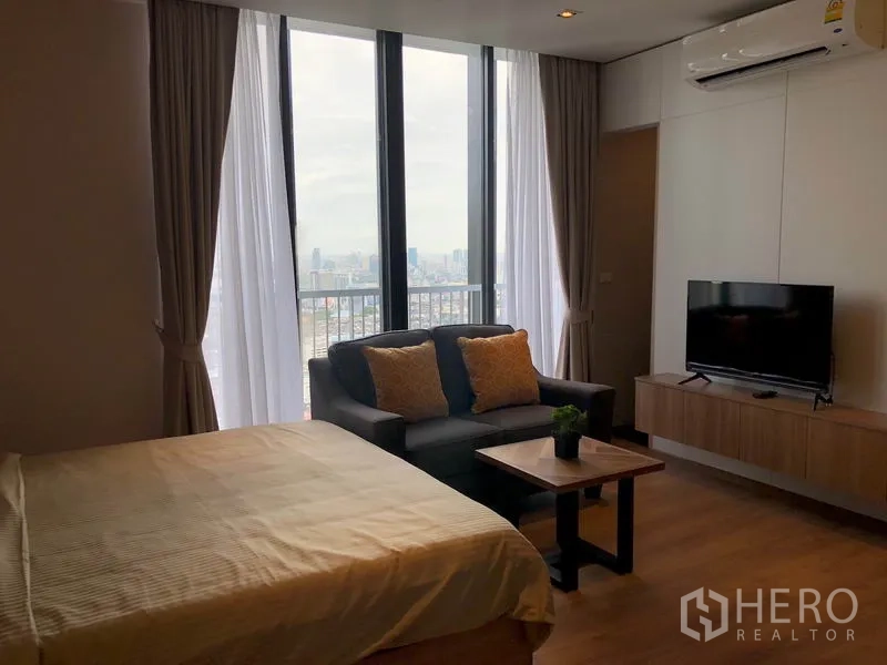 Condominium for rent in Khlong Toei, Bangkok - Room with city view through tall windows, sofa set and wall-mounted TV beside the bed.