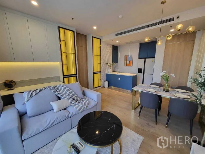 Condominium for rent in Watthana, Bangkok - Bright open‑plan living area with pastel sofa, black marble coffee table, blue kitchen island and dining set under brass pendant lights.