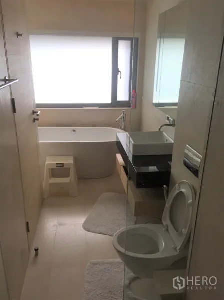 Condominium for rent in Bang Rak, Bangkok - Bathroom featuring a bathtub below a frosted window, wall-mounted sink and toilet.
