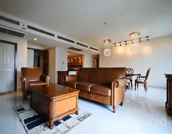 公寓 出租在Pathum Wan, Bangkok - 采光充足的客厅，棕色真皮沙发、木质茶几与水晶吊灯，旁接餐区。