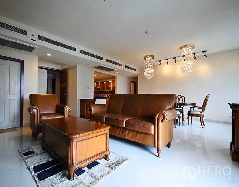 คอนโด ให้เช่าในPathum Wan, Bangkok - ห้องนั่งเล่นโปร่งสว่าง ชุดโซฟาหนังสีน้ำตาล โต๊ะกลางไม้ และโคมไฟคริสตัลข้างโซนรับประทานอาหาร