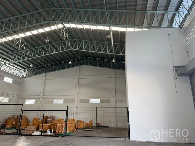 Warehouse for rent in Lat Krabang, Bangkok - High-clear warehouse roof trusses, white service core, and fenced zone with packed goods.
