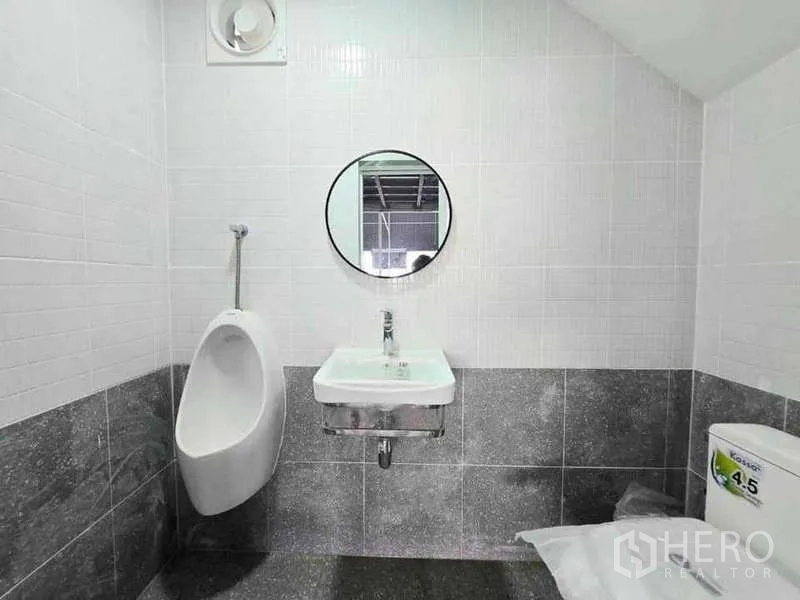 Warehouse for rent in Prawet, Bangkok - Clean tiled restroom with urinal, sink, round mirror and toilet inside the warehouse.