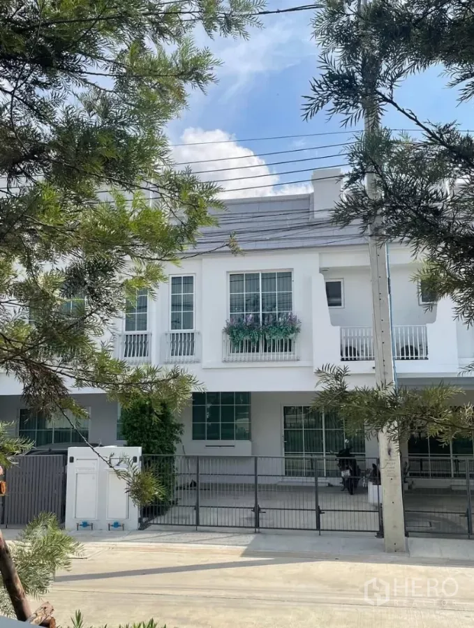 Townhouse for rent in Bang Phli, Samut Prakan - Two‑storey white townhouse facade with gated parking and balcony planter framed by trees.