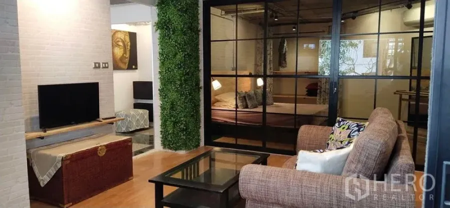 Condominium for sale in Watthana, Bangkok - Open-plan living and bedroom divided by black-framed glass, TV console, sofa and vertical garden wall.