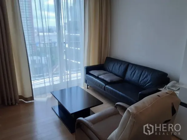 Condominium for rent in Khlong Toei, Bangkok - Bright living area with black leather sofa, armchair and coffee table opening to a balcony with city view through sheer curtains.